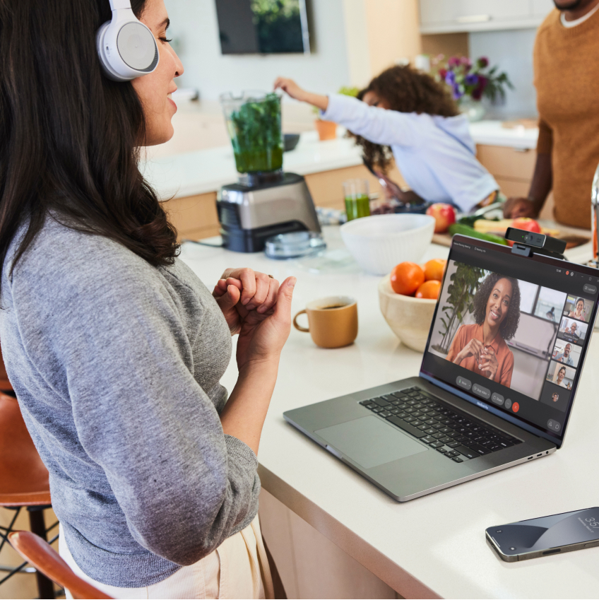 A person wearing a headset engages in a Webex Meeting as family members make juice in a blender.