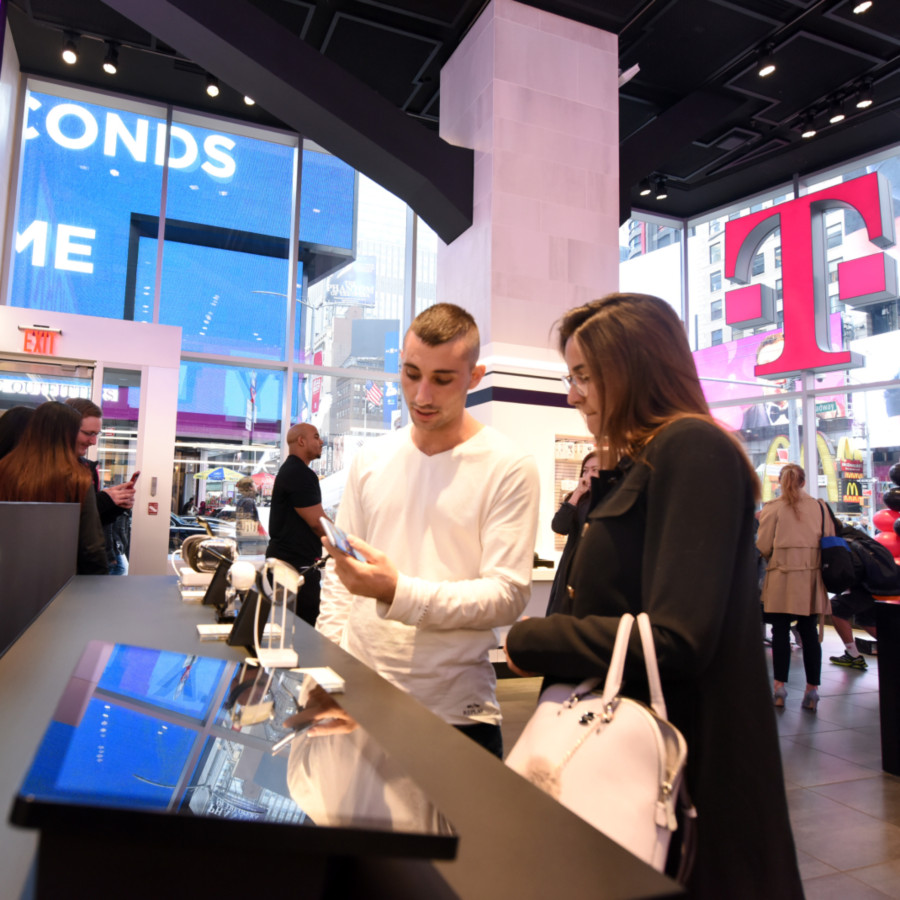 Two people look at a mobile phone in a large, bustling room with a giant pink T-Mobile logo on the wall behind them.