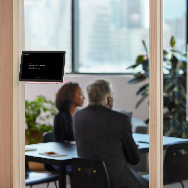 A Webex Room Navigator glows red outside a conference room where two colleagues are meeting, indicating the room is booked.