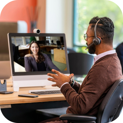 A person wearing a headset with a microphone talks to another person via a monitor and webcam.