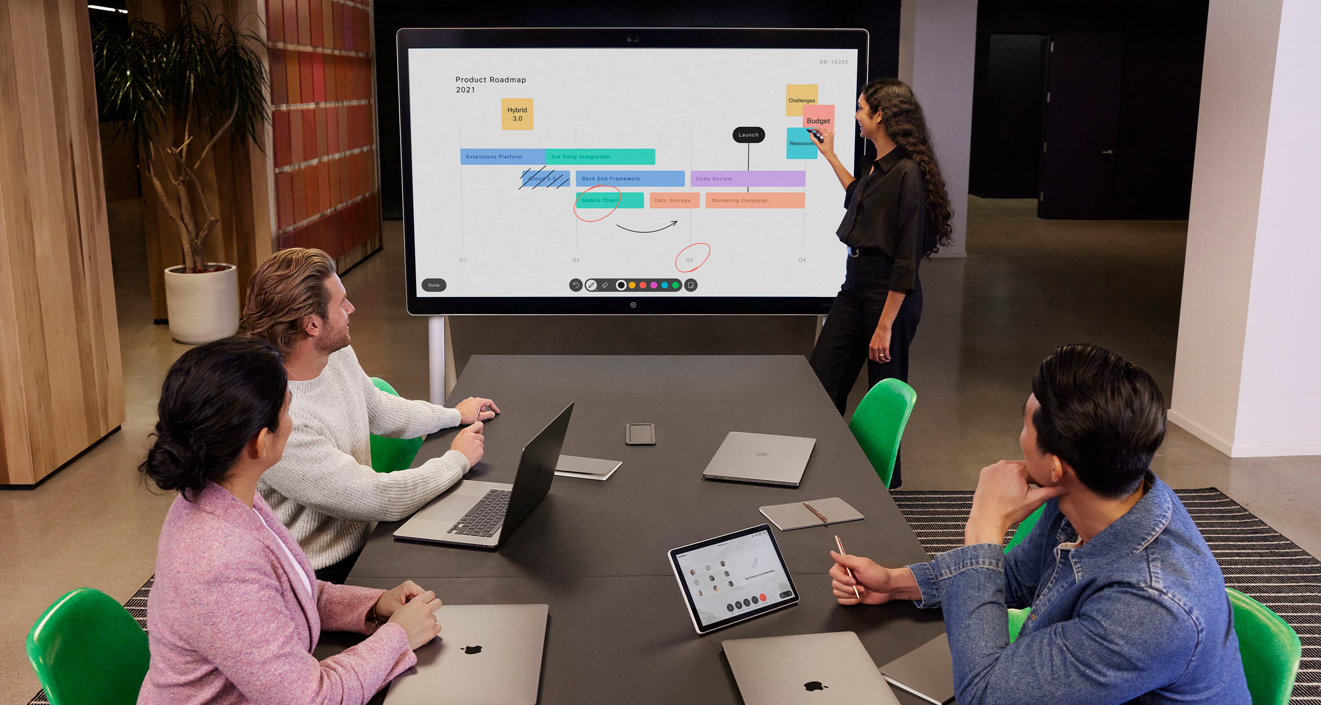 Three colleagues sit at a table in a meeting room, listening as one colleague talks at the front. Everyone has MacBooks.