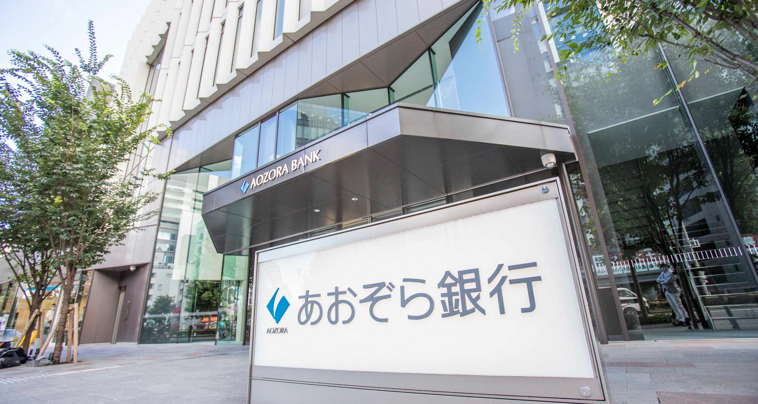 The exterior of the Aozora Bank office on a tree-lined street. The name Aozora Bank is visible above the glass office doors.