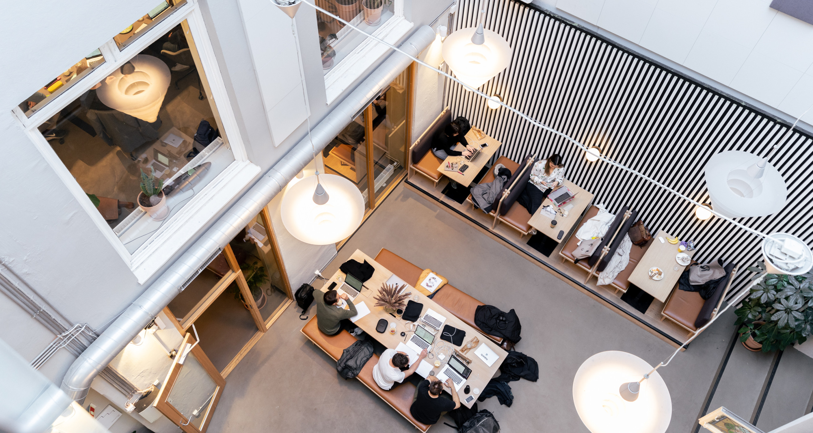 People work in a modern open workspace with high ceilings, lots of light, 3 smaller tables, and a long table in the center.