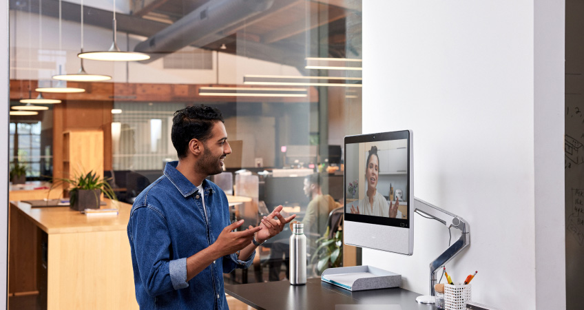 Person in a denim button down shirt at a standing desk in an office collaborates with a colleague via a video meeting.
