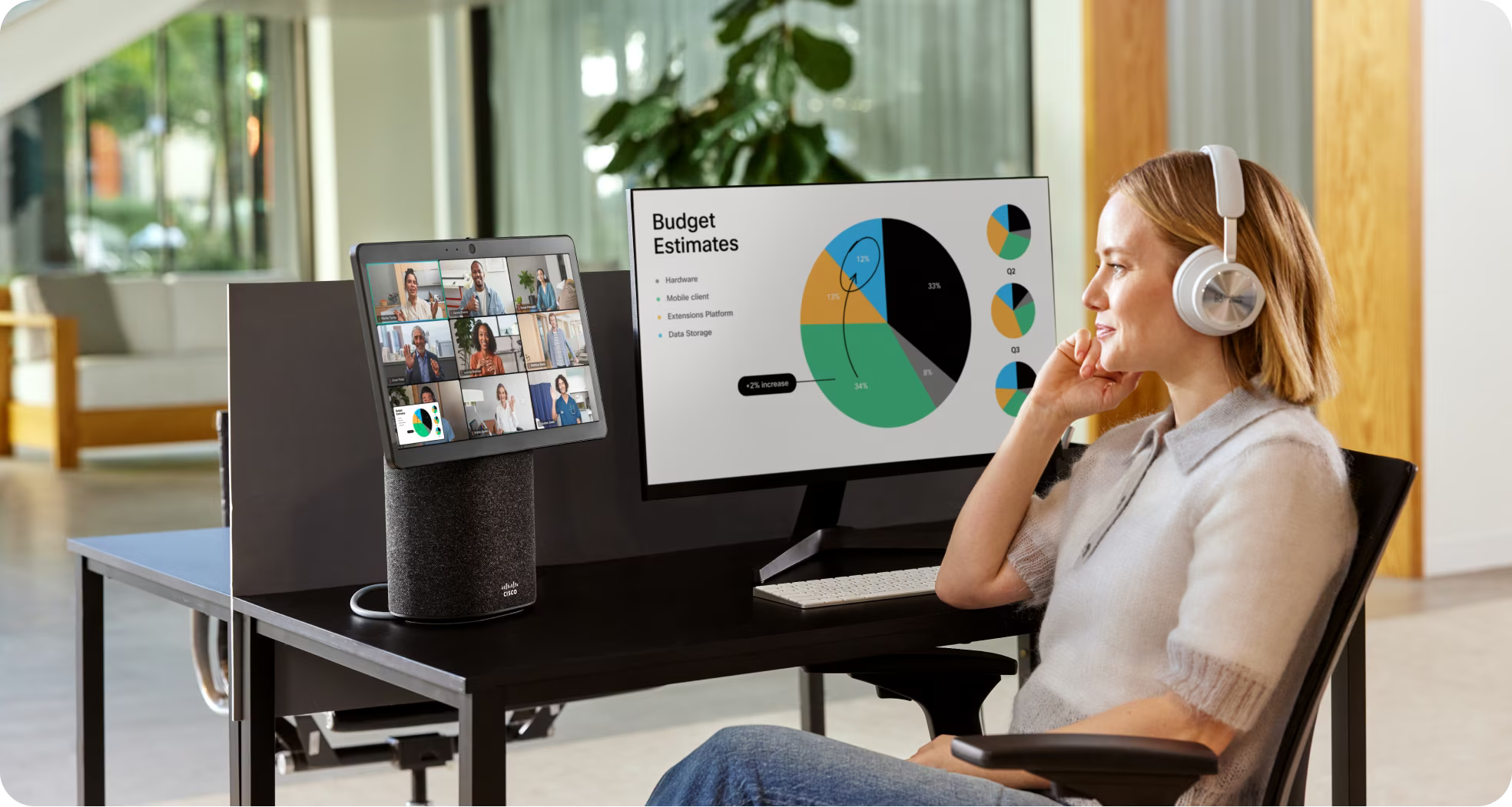 Person in a headset video conferences with colleagues on a Webex Desk Mini. Her second monitor shows a Budget Estimate slide.