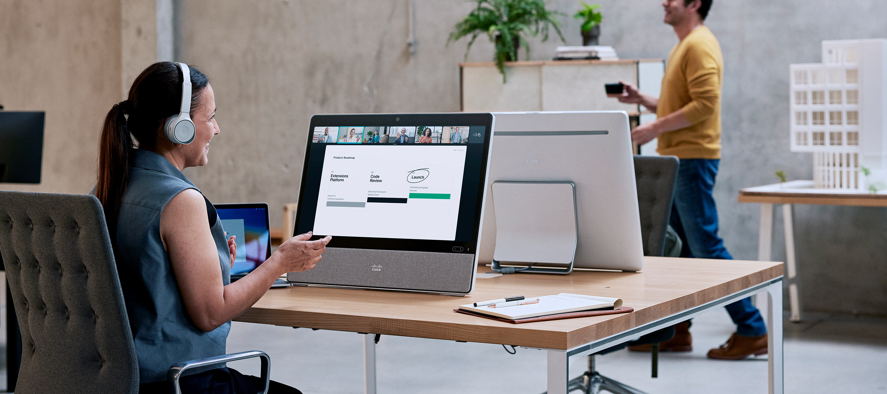 Sitting at a desk in an open office, a professional wearing a headset video conferences with a colleague.